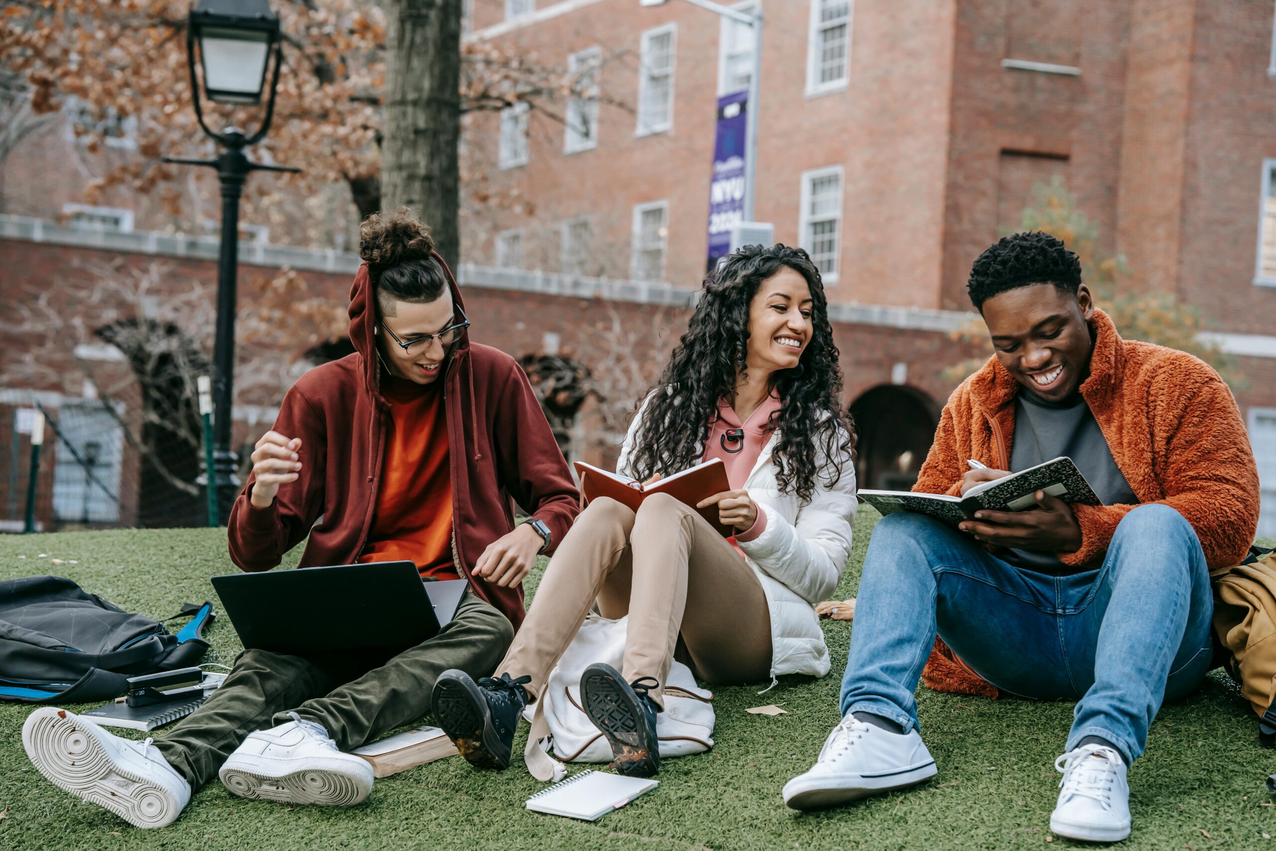 Home Full body of happy diverse students with notebooks and laptop sitting on grassy lawn on campus of university while studying together